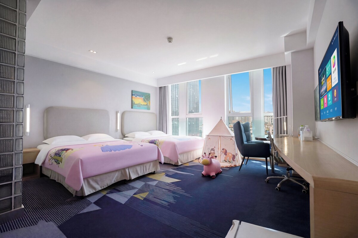 A hotel room with two beds, a desk, a chair, a TV, and large windows.