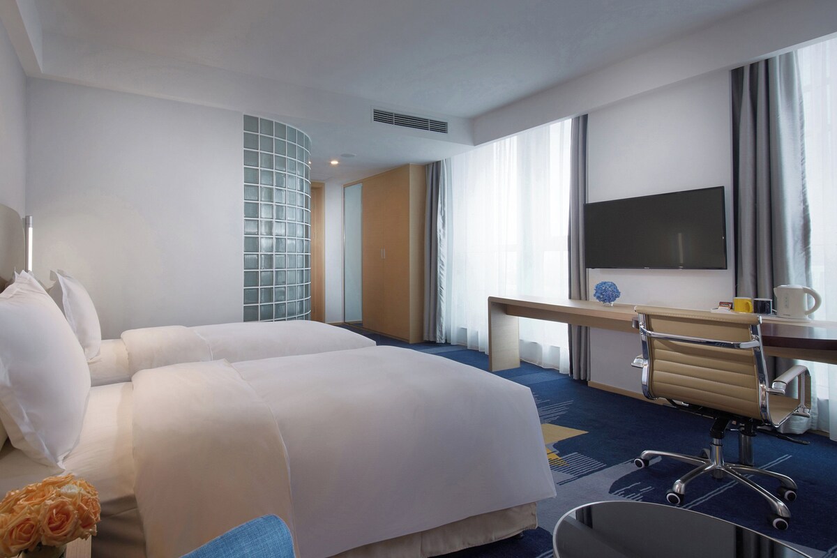 A hotel room with a large bed, a desk, a chair, a TV, and a window with curtains.