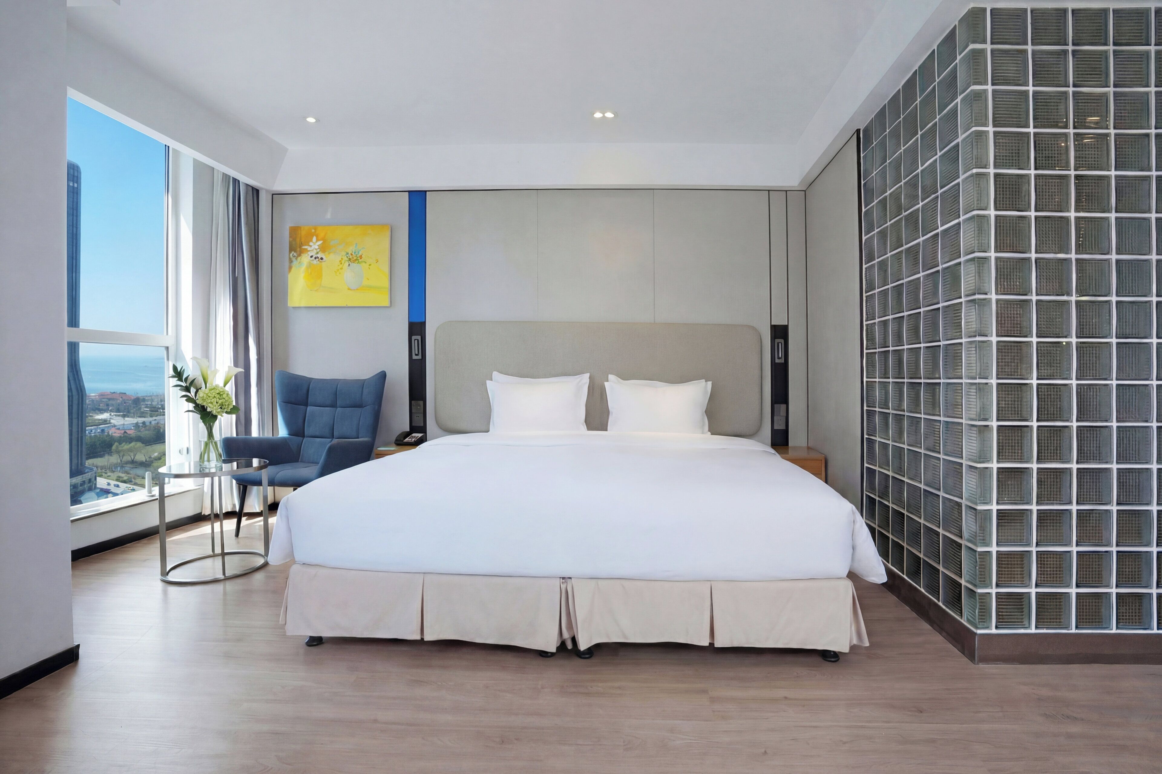A modern hotel room with a large bed, a chair, a small table, and a city view.