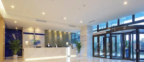 Un hall d’hôtel moderne avec un comptoir d’accueil, une grande porte d’entrée en verre et un sol en marbre.