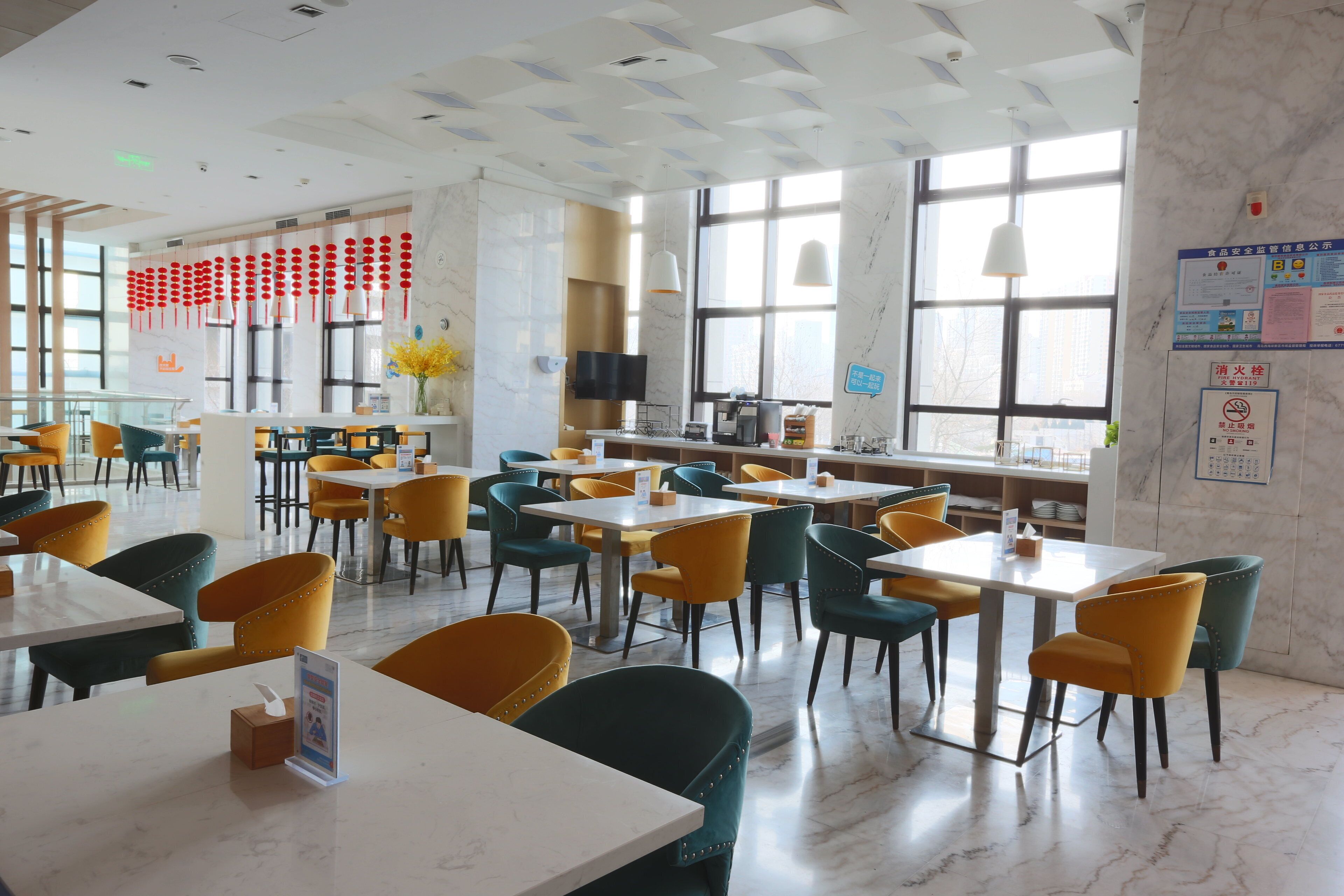 A spacious dining area with multiple tables and chairs, large windows, and a modern ceiling design.