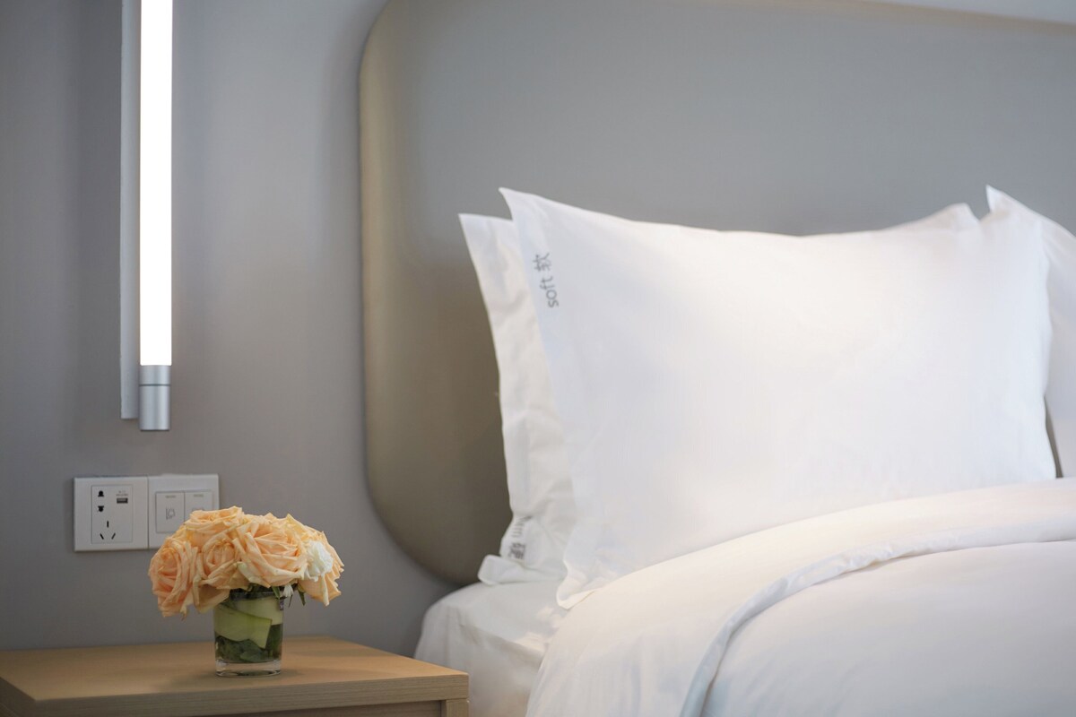 A neatly made bed with white linens and pillows, a bedside table with a vase of flowers, and a wall-mounted light fixture.