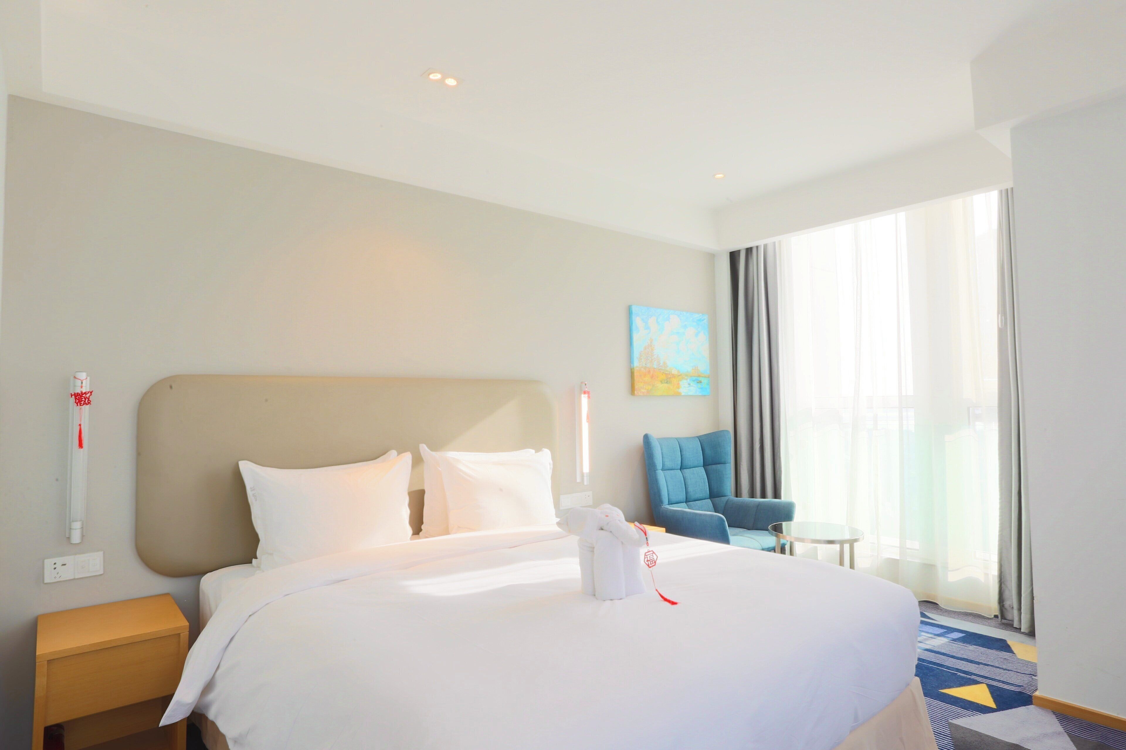 A hotel room with a large bed, a nightstand, a blue armchair, a small table, and a large window with curtains.