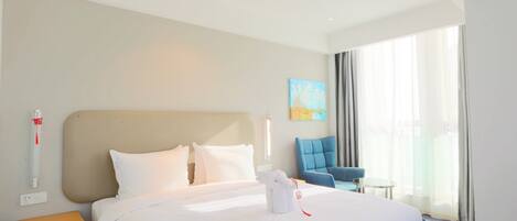 A hotel room with a large bed, a nightstand, a blue armchair, a small table, and a large window with curtains.