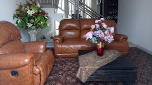 A living room with a brown leather sofa, a coffee table with a vase of flowers, and a patterned rug. A staircase is visible in the background. - T-Rooms Homestay (Palembang)