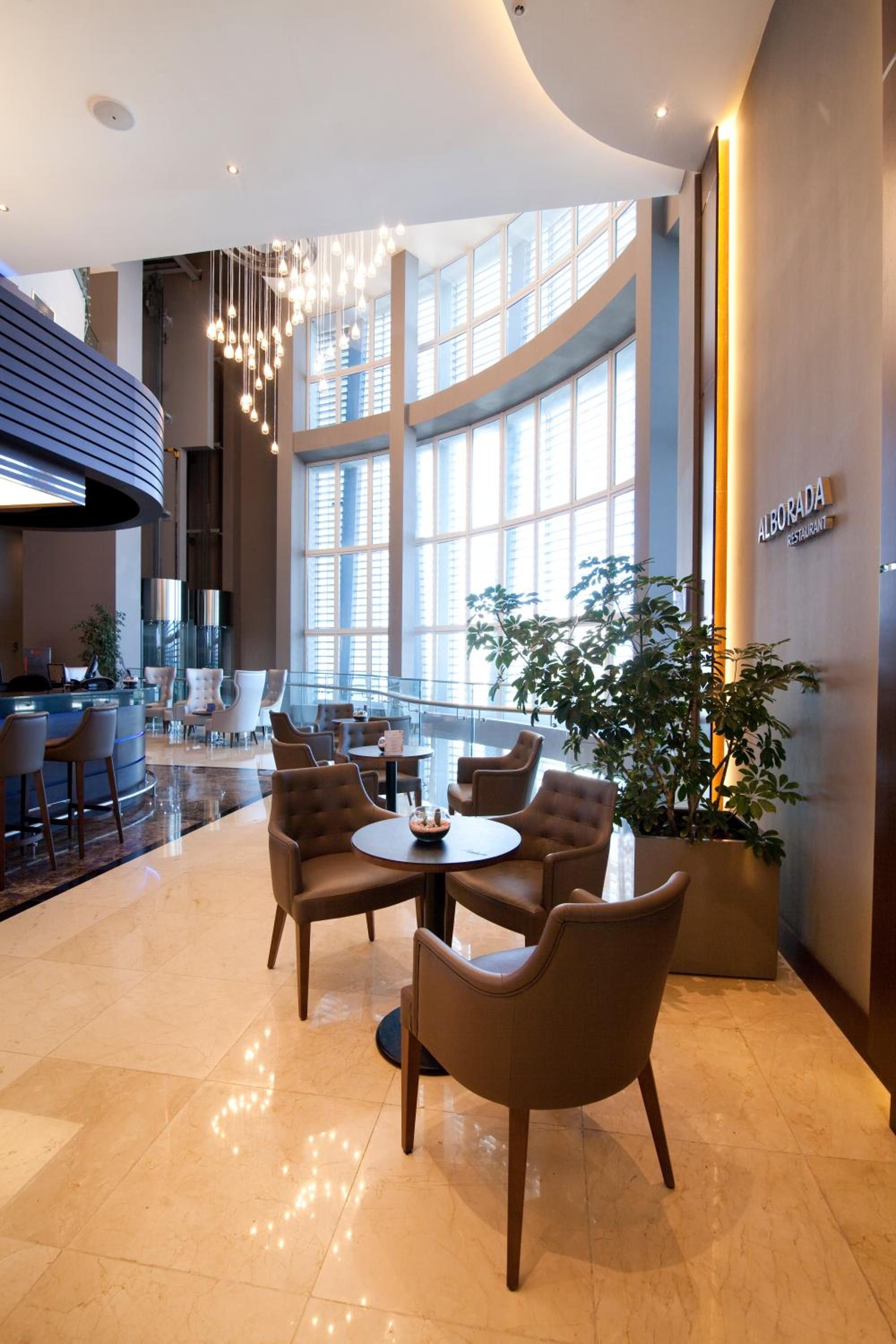 A spacious indoor area with a central round table surrounded by chairs, large windows, and a sign for 'Alborada Restaurant'.