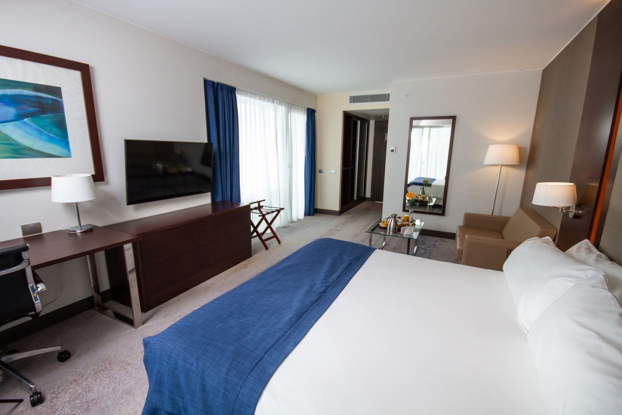 Ein Hotelzimmer mit einem großen Bett, einem Schreibtisch mit Stuhl, einem Fernseher, einem Spiegel und einer Lampe.
