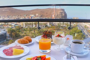 Una mesa de desayuno con croissants, fruta y bebidas, con vistas a una ciudad costera.