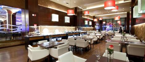 Restaurante moderno com várias mesas de jantar, cadeiras brancas e iluminação pendente vermelha.