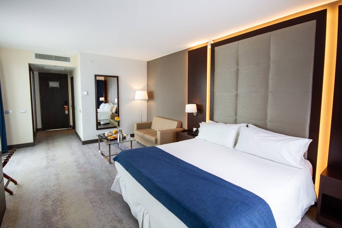 A hotel room with a large bed, a sofa, a small table with a lamp, and a mirror.