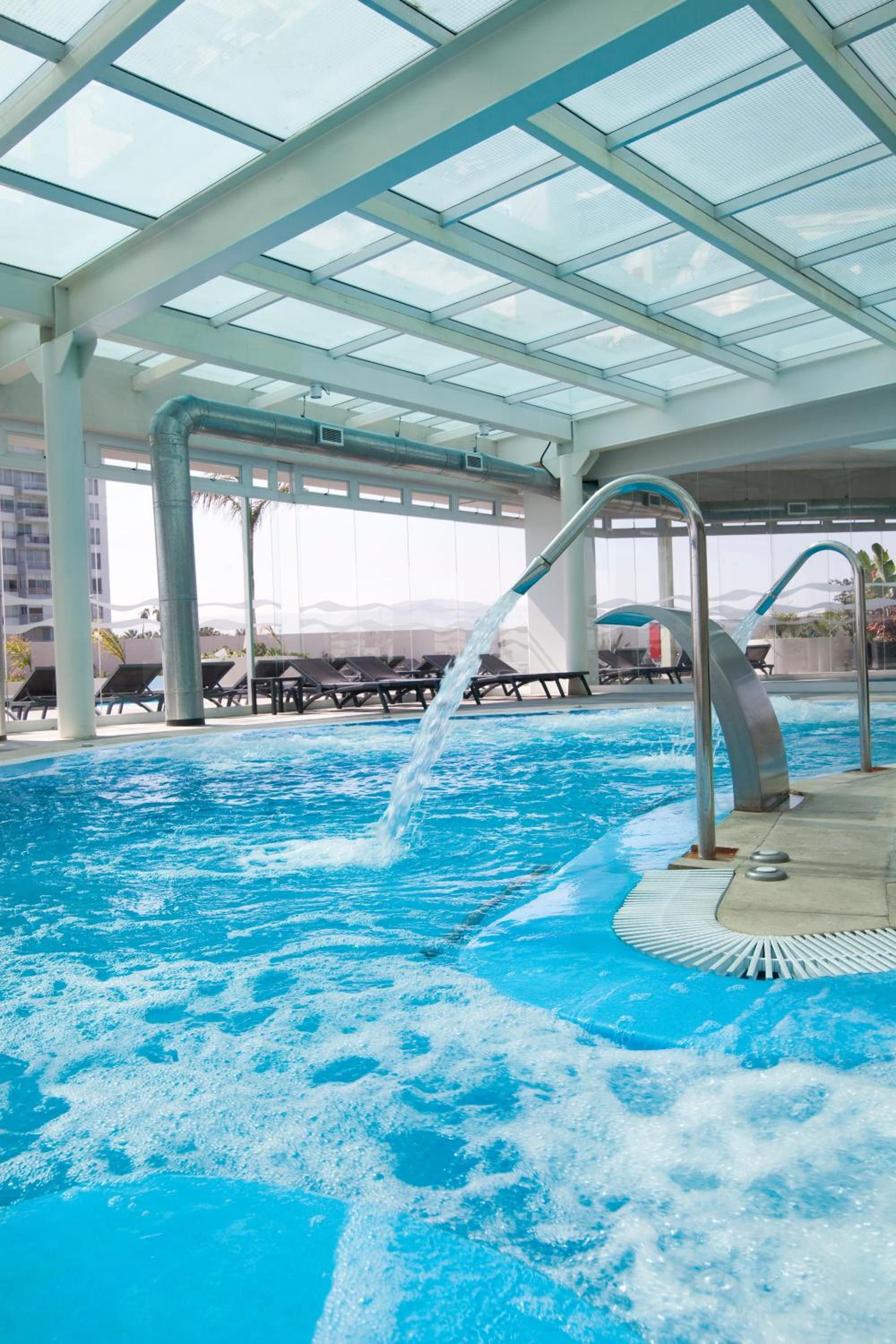 An indoor swimming pool with clear glass ceilings, lounge chairs, and a water feature.