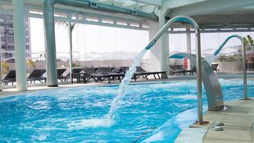 An indoor swimming pool with clear glass ceilings, lounge chairs, and a water feature.