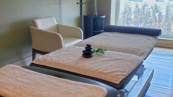 Una habitación con dos camillas de masaje, una silla, una pequeña planta en maceta y una ventana con vistas a árboles.