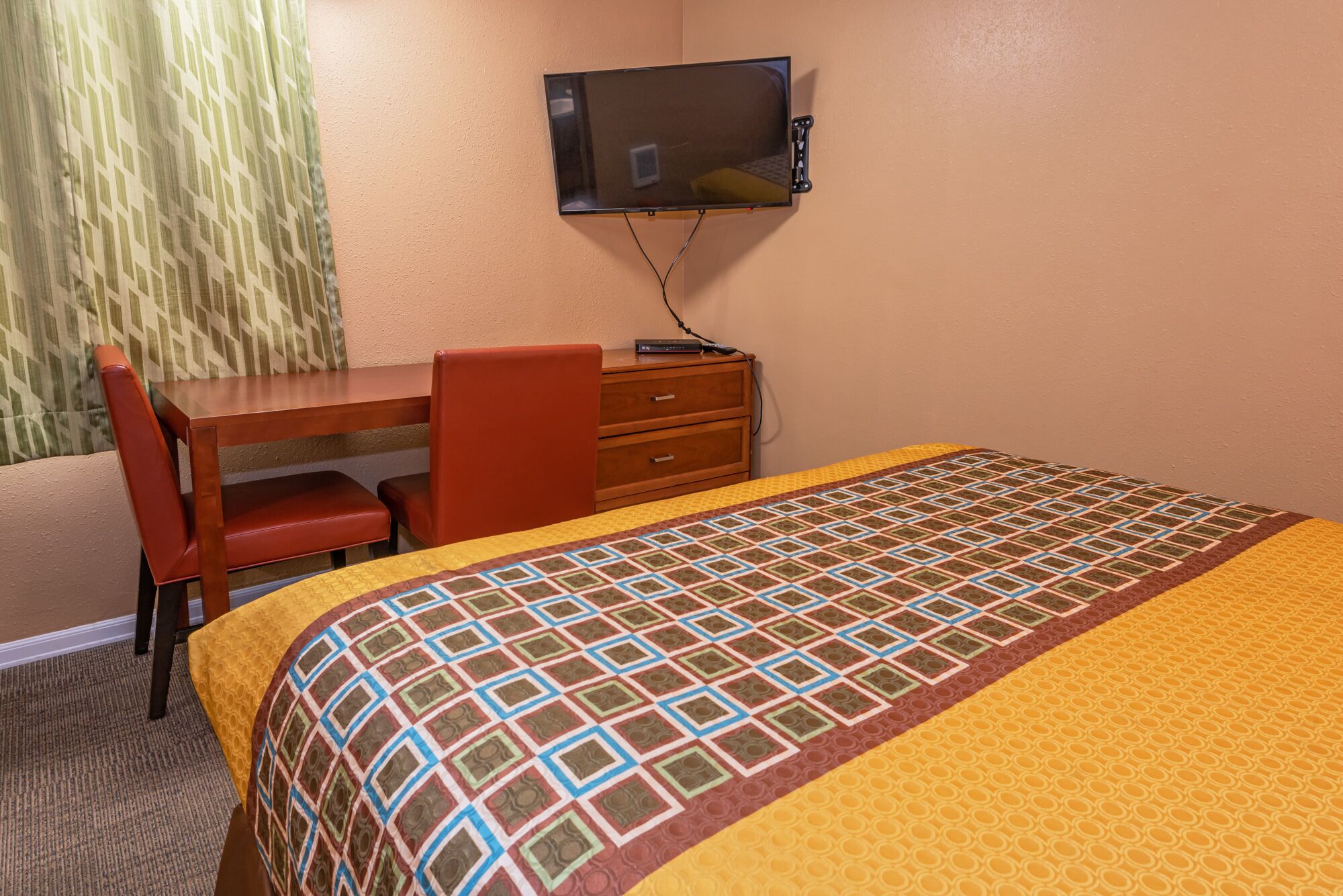 Habitación de hotel con cama, escritorio, silla, televisor montado en la pared y ventana con cortinas.