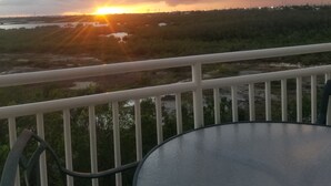 Un balcón con una mesa redonda y sillas, con vista a una puesta de sol sobre un paisaje pantanoso.
