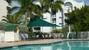 Una piscina con sillones de descanso y una zona sombreada, rodeada de palmeras y un edificio con balcones.
