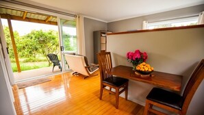 Una habitación con piso de madera, una mesa de comedor con sillas, una mesita auxiliar con un florero y un puerta corrediza de vidrio que da a un espacio exterior con vegetación.