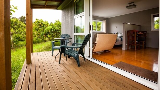 Una terraza de madera con una mesa y sillas, una puerta corrediza de vidrio que da a un dormitorio y vistas al exterior con vegetación.