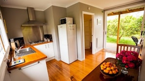 Cocina compacta con fregadero, estufa y electrodomésticos blancos. Se ve una mesa de comedor con un cuenco de frutas y flores.