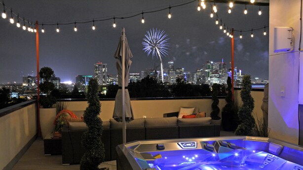 Una terraza en la azotea con bañera de hidromasaje, luces colgantes y un paisaje urbano con fuegos artificiales al fondo.