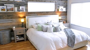 Un dormitorio con pared de madera, una cama con ropa de cama blanca, dos mesitas de noche con lámparas y una ventana con persianas.