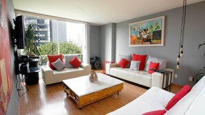 A modern living room with two white sofas, red and white cushions, a wooden coffee table, and a large window offering a view of buildings and greenery. - Prime Roma Norte 2BR; Walk to Cibeles, Reforma & Condesa (México D.F.)