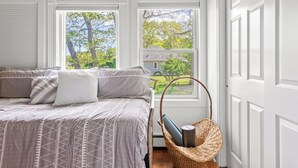 Une chambre à coucher avec un lit, deux fenêtres, un fauteuil en osier et une vue sur une maison.
