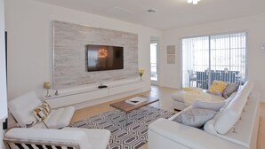 Un soggiorno moderno con una grande TV a schermo piatto montata su una parete di legno chiaro, un divano bianco, un tappeto decorato e un tavolino da caffè.