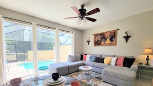 A living room with a grey sofa, a ceiling fan, a large window, and a pool view.
