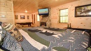 Un acogedor interior de cabaña con chimenea de piedra, televisor de pantalla plana montado en una pared de piedra y una alfombra grande con estampado.