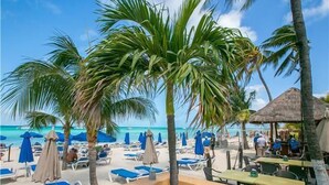 A beach with lounge chairs and umbrellas, palm trees, and a thatched-roof structure. - Nautibeach Condo 22  – 2 Bedroom, Family-Friendly Vacation on the Beachfront Third-Floor (Isla Mujeres)