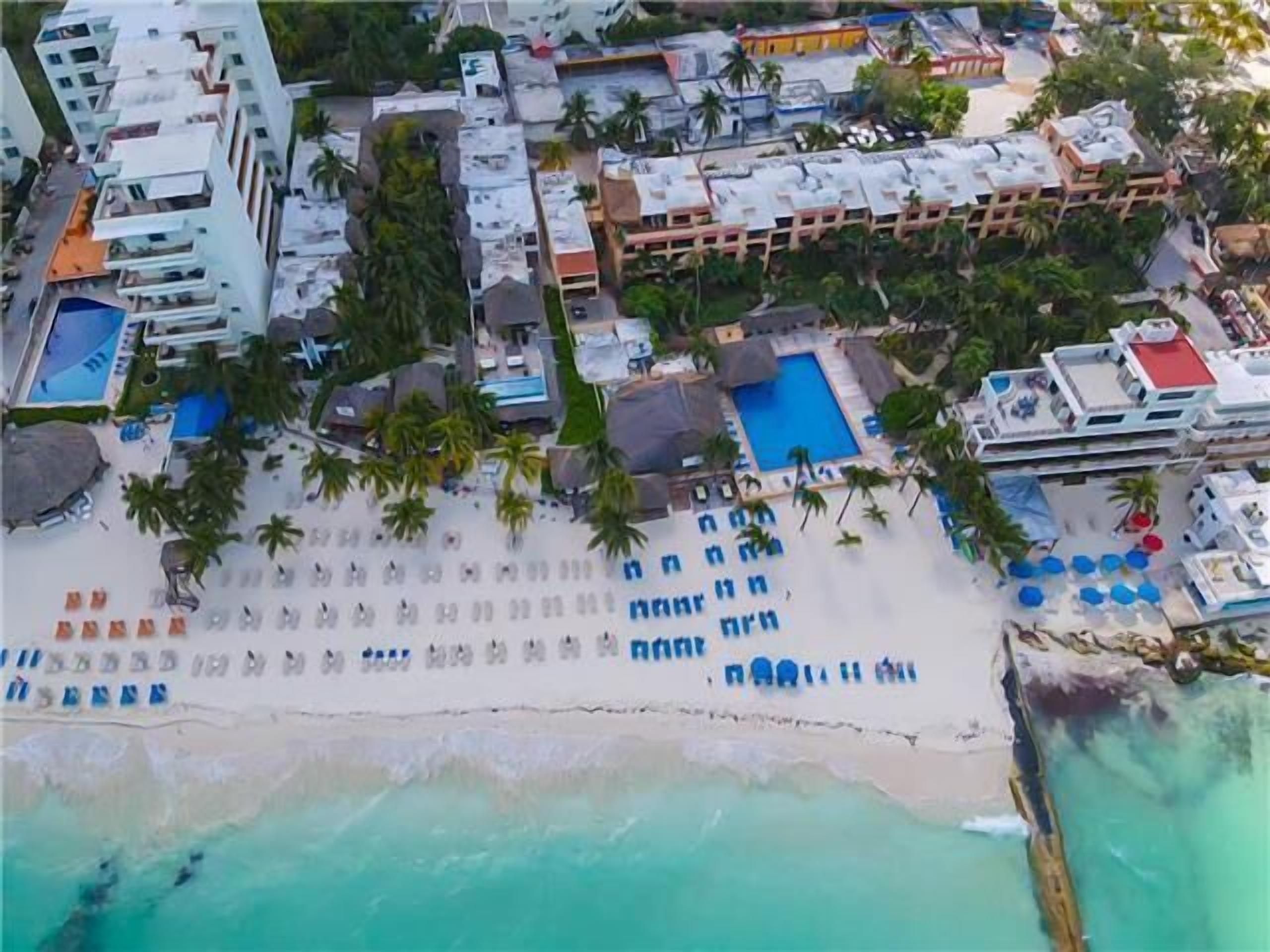 Vue aérienne d’un complexe hôtelier comprenant plusieurs bâtiments, une plage avec des transats et une piscine.