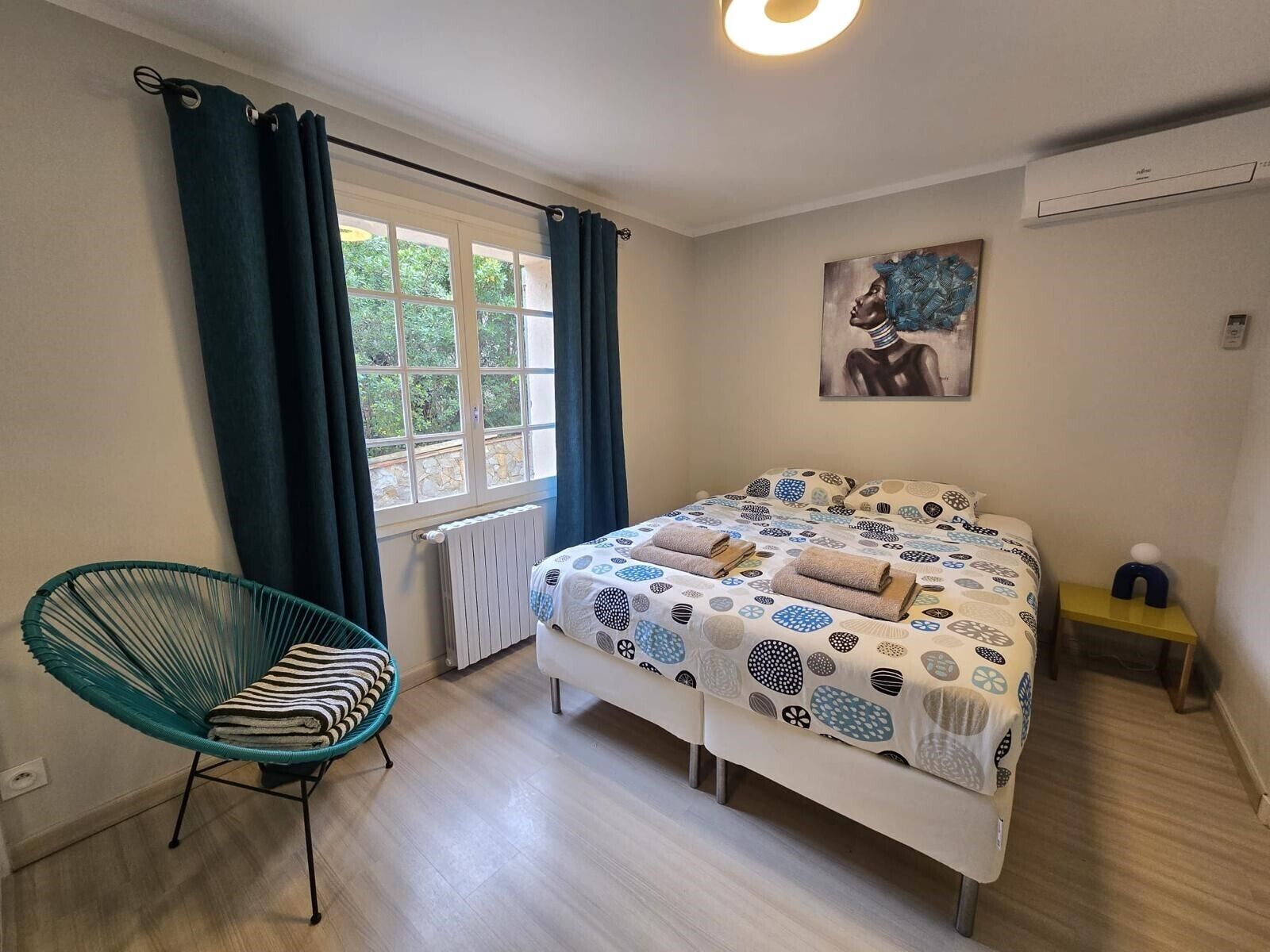 Un dormitorio con cama, una silla, un radiador, una ventana con cortinas, un cuadro y una mesita de noche.