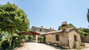 Un edificio rustico in pietra con tetto in tegole, un pergolato in legno e una strada sterrata che conduce a una casa con tetto in tegole.