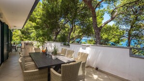 A covered outdoor dining area with wicker furniture, a table set with glasses and a bottle, and a view of trees and a body of water. - Beachfront apartment with Infinity Pool & BBQ (Slatine)