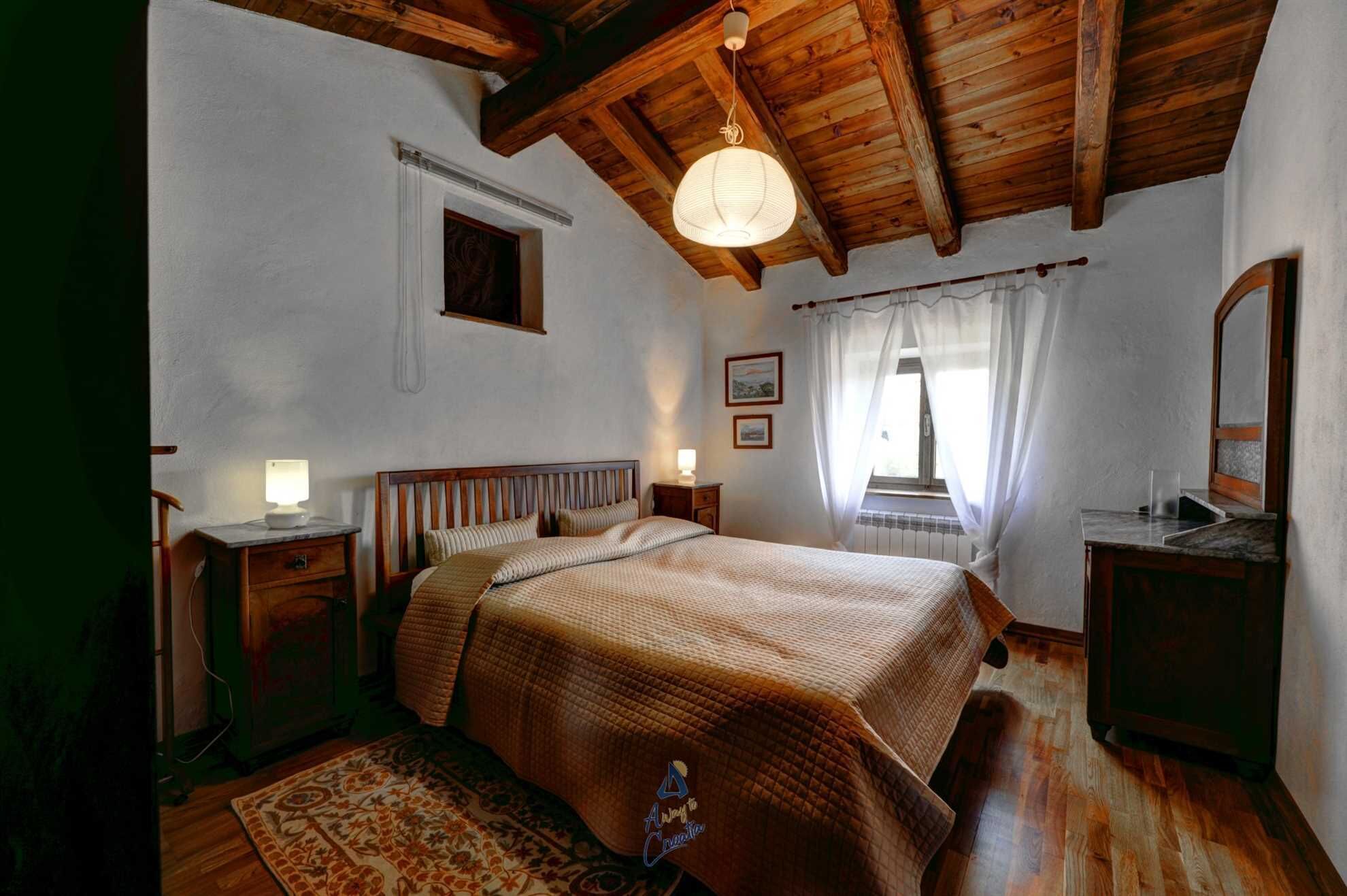 Une chambre avec un plafond en bois, un lit recouvert d’une couverture matelassée, une table de chevet avec une lampe, une fenêtre avec des rideaux et un miroir sur le mur.
