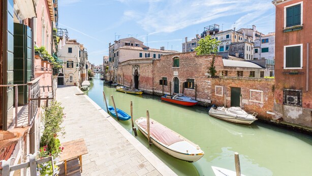 A narrow canal with boats docked along the sides, surrounded by historic buildings with visible architecture and a clear sky. - Ca' Tintoretto (Venice)