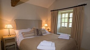 A neatly made bed with a beige bedspread and pillows, two bedside lamps, and a window with curtains. - Luxurious Converted Bothy/Barn In an Idyllic Setting (West Burton)