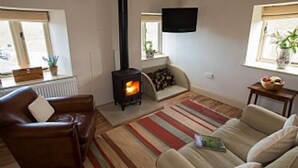 A cozy living room with a wood-burning stove, leather sofas, a TV mounted on the wall, and a window with a plant. - Luxurious Converted Bothy/Barn In an Idyllic Setting (West Burton)