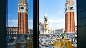 Uma varanda com toalha de mesa amarela, um vaso decorativo e vista para duas torres imponentes, com um campanário ao fundo.