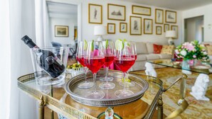 Uma bandeja com copos de vinho e uma garrafa sobre uma mesa em uma sala de estar, com um sofá, quadros e objetos decorativos.