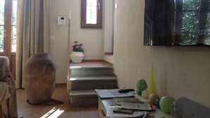 Una sala de estar con un televisor montado en la pared, un jarrón grande con flores, una escalera y una ventana con cortinas.