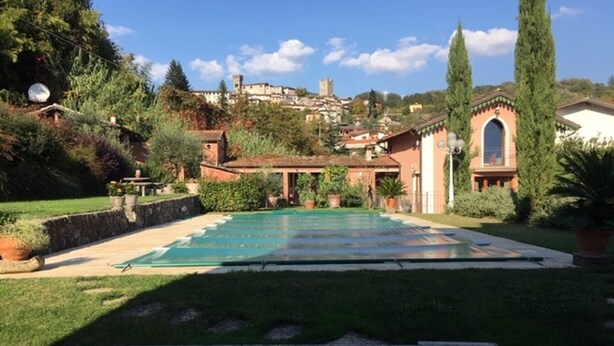 Piscina rodeada de jardín, barandilla de piedra y vista a una colina con edificios.