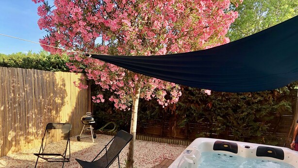 Un pati amb una banyera d'hidromassatge, un pont de fusta, un barbacoa i un arbre amb flors roses.