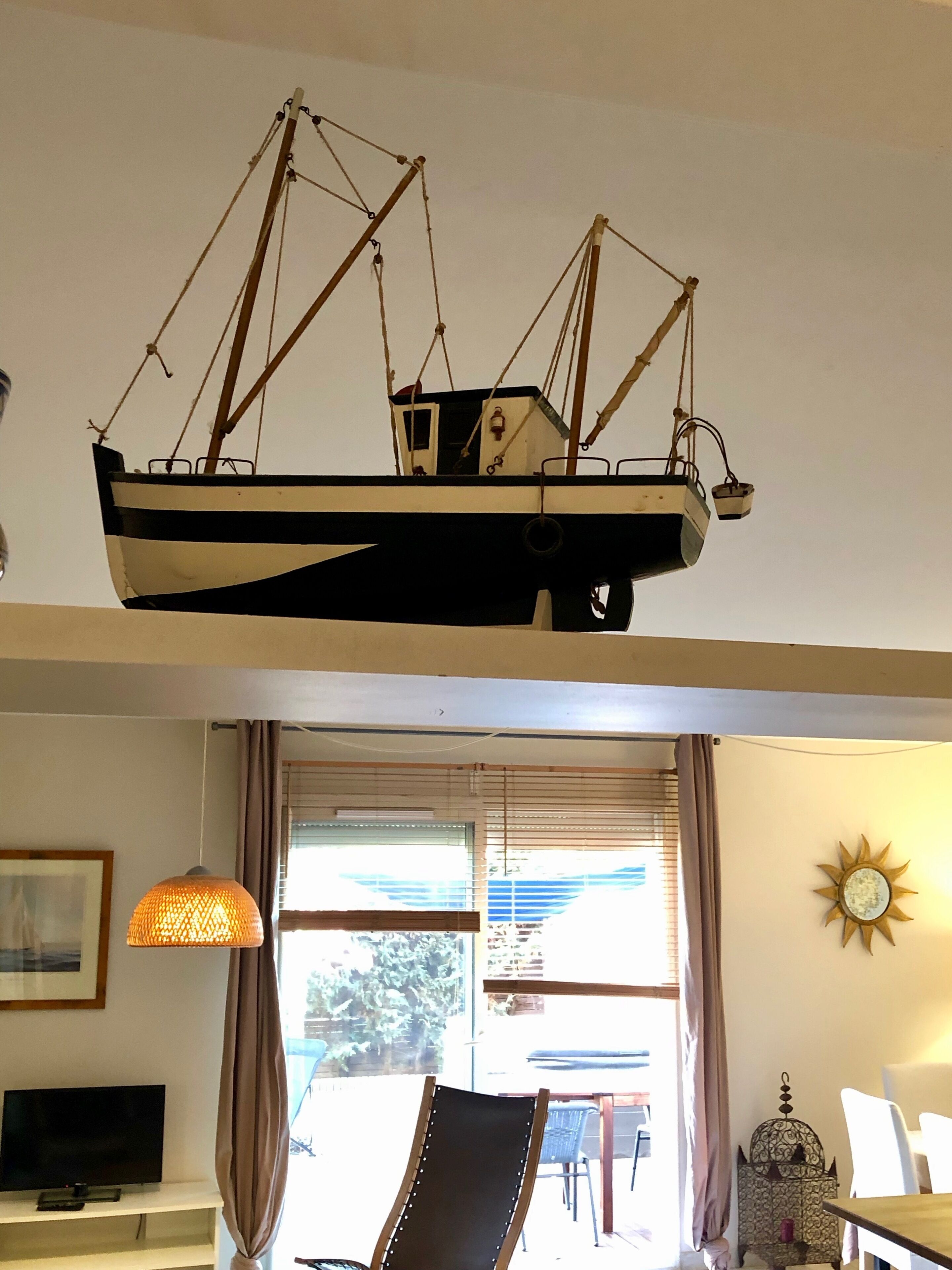 Une pièce comprenant un téléviseur posé sur un meuble, une chaise, une table, un luminaire suspendu et une maquette de bateau décorative.