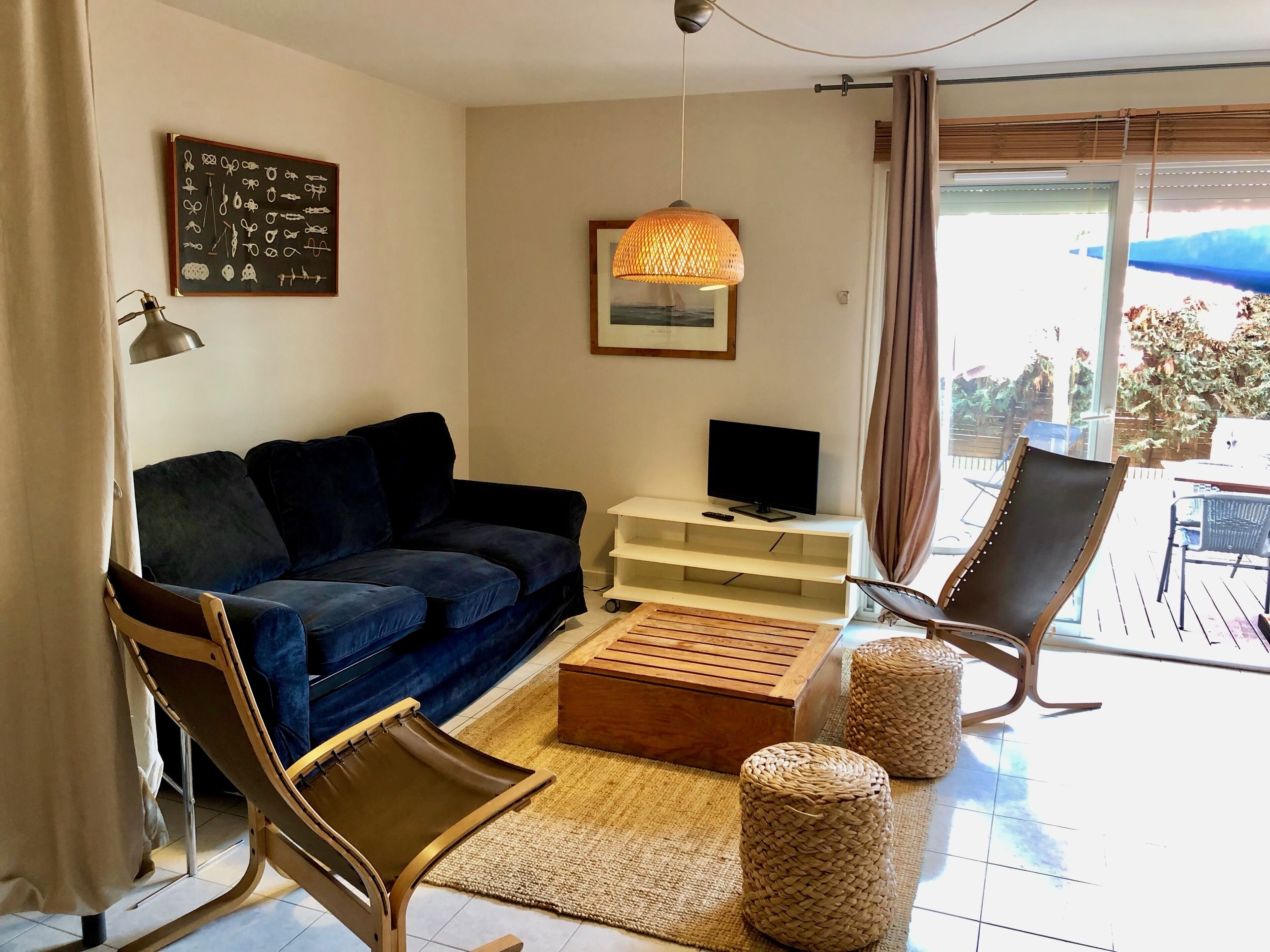 Un salon avec un canapé bleu foncé, une table basse en bois, un pouf en osier, une chaise en osier, un téléviseur posé sur un meuble blanc et un luminaire suspendu.