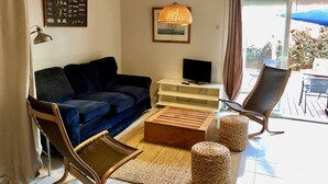 Un saló amb un sofà blau fosc, una taula de centre de fusta, un otomà de vímet, una cadira de vímet, un televisor sobre un prestatge blanc i una làmpada penjant.