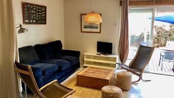 Un salon avec un canapé bleu foncé, une table basse en bois, un pouf en osier, une chaise en osier, un téléviseur posé sur un meuble blanc et un luminaire suspendu.