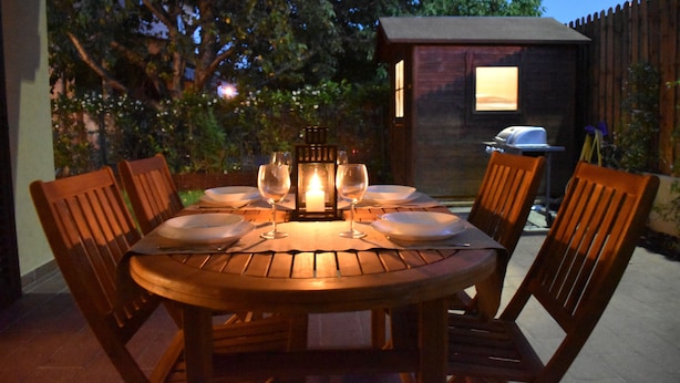 An outdoor wooden table set for dining with chairs, a lit lantern, and a view of a backyard with a barbecue grill. - Seaside house with private garden (Follonica)