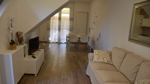 A living room with a sofa, a TV, a dining table with chairs, and a staircase leading to an upper level. - Seaside house with private garden (Follonica)